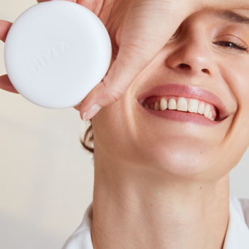Okrągły, biały podkład NIVEA Cellular 3in1 w kompakcie, trzymany blisko twarzy uśmiechniętej kobiety.