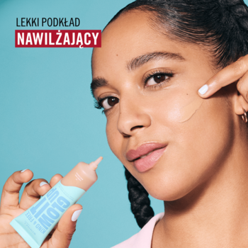 Kobieta o ciemnej karnacji nakłada nawilżający podkład Rimmel Glow It Up z niebieskiej tubki na policzek. Tło błękitne.