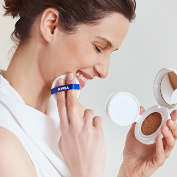 Kobieta aplikuje podkład NIVEA Cellular Care Cushion 3w1, widać otwarte opakowanie i biały puff NIVEA.
