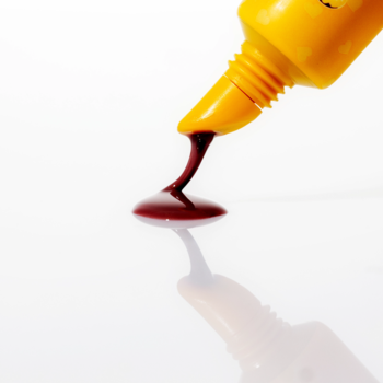 Fluff Candy Lip Balm Vanilla Glazed Donut: żółta tubka wyciskająca brązowy balsam na błyszczącej, białej powierzchni.