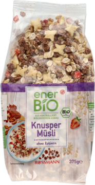 Przezroczysta torba enerBiO Knusper Müsli z widocznymi kawałkami kakaowego crunchy, żółtymi gwiazdkami i truskawkami.