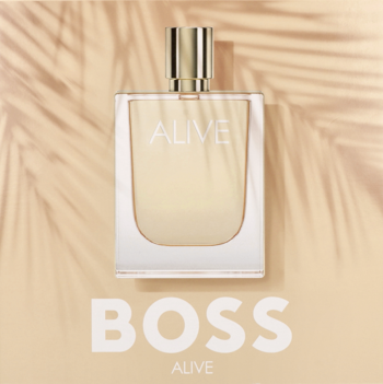 Elegancka butelka perfum HUGO BOSS Alive z złotym korkiem na jasnym tle z cieniami liści palmowych.