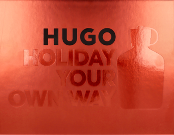 Czerwone tło z czarnym napisem HUGO, wytłoczony tekst "HOLIDAY YOUR OWN WAY" i zarys butelki Hugo Man.