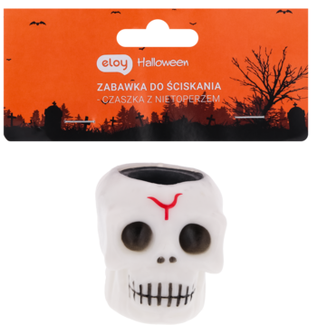 Biała zabawka-gniotek czaszka z czarnymi oczami i czerwonym symbolem, przed pomarańczowym opakowaniem Halloween.