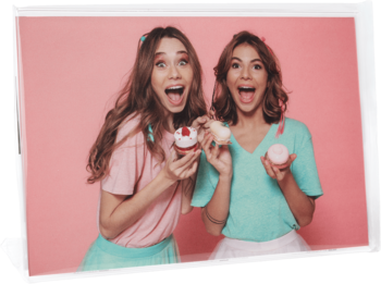 Dwie roześmiane młode kobiety w pastelowych ubraniach trzymają kolorowe babeczki z kremem na różowym tle.