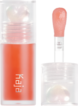 Kaja Juicy Glass Lip Oil Apricot Allure. Pomarańczowy olejek do ust w butelce z sercem, obok wyjęty aplikator z produktem.
