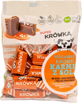 Biała torebka Super Krówka: wegańskie krówki o smaku karmelu z solą, z rozpakowanymi cukierkami obok.