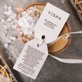 Biała zawieszka Yesha Mocca Cafe do soli do kąpieli, leżąca na soli i drewnie, widok z góry.