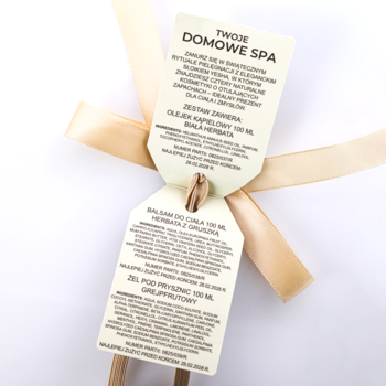 Dwie białe etykiety prezentowe zestawu SPA (olejek, balsam, żel) przewiązane beżową wstążką, widok z góry.