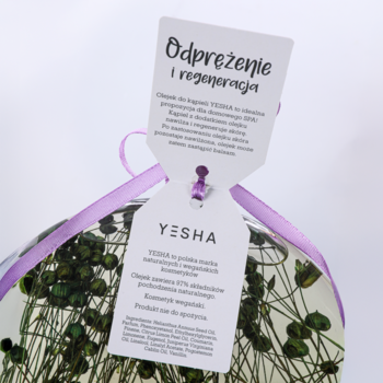 Olejek do kąpieli Yesha Safari w przezroczystym pojemniku z zielonymi roślinami, z białą metką i fioletową wstążką.