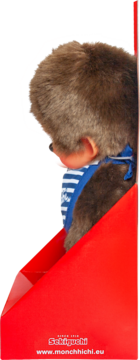 Tył pluszowej zabawki Monchhichi z brązowym futrem, w niebieskim śliniaku, w czerwonym pudełku ekspozycyjnym.