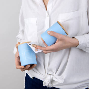 Kobieta trzyma dwie jasnoniebieskie świece Dorota & Aroma Home z drewnianymi wieczkami i widoczną metką produktu.