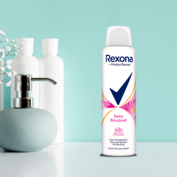 Rexona Sexy Bouquet antyperspirant w sprayu, biała butelka z różowo-pomarańczowymi kwiatami, na tle łazienki.