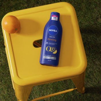 Nivea Q10 Firming + Witamina C mleczko do ciała, granatowa butelka na żółtym stołku obok pomarańczy, zielona trawa.