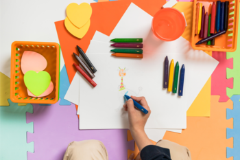 Trójkątne kredki świecowe w akcji: dziecko rysuje niebieską kredką, wokół żywe kolory i akcesoria na macie.