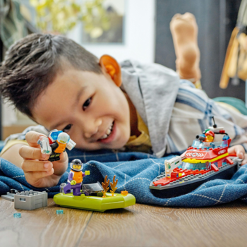 Uśmiechnięty chłopiec leży na podłodze, bawiąc się zestawem LEGO z czerwoną łodzią i minifigurkami ratowników.
