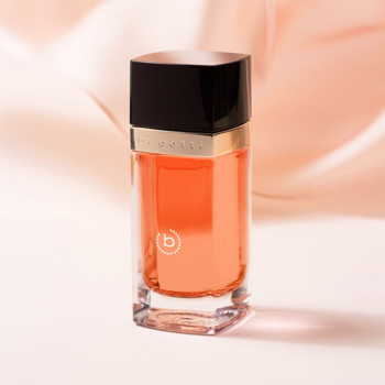 Bugatti Eleganza Ambra woda perfumowana. Pomarańczowa butelka z czarną nakrętką, logo na złotej listwie, beżowe tło.