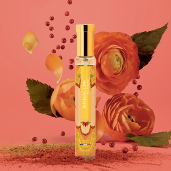 Perfumy Adopt Bombay Chic, żółta butelka rollerball otoczona kwiatami i różowym pieprzem na koralowym tle.