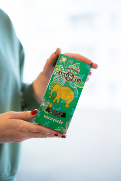 Zielone pudełko herbaty Adalbert's Tea Green Fruits SZCZĘŚCIE z żółtym słoniem, trzymane w dłoniach.