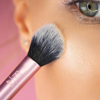 Zbliżenie pędzla Real Techniques Makeup Setting Brush z różową rączką, aplikującego makijaż na policzek.