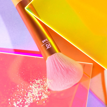 Zbliżenie na pędzel Real Techniques Glow Off Multitasking Brush z różowym włosiem dotykającym sypkiego pudru, na kolorowym tle.