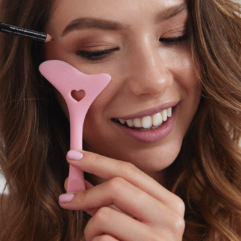 Uśmiechnięta kobieta używa różowego silikonowego szablonu do makijażu oczu z sercem, do precyzyjnych kresek.