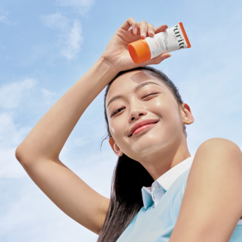 Uśmiechnięta kobieta trzyma nad głową pomarańczowo-biały krem przeciwsłoneczny SPF 50+ pod słońcem.