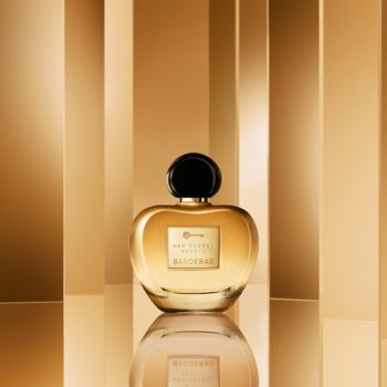 Antonio Banderas Her Secret Absolu, złoty flakon perfum o opływowym kształcie z czarną nakrętką na tle złotych paneli.