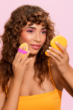 Kobieta z kręconymi włosami używa żółto-fioletowego aplikatora Miracle Powder Puff, patrząc w lusterko.