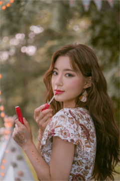 Kobieta nakłada Rom&nd Juicy Lasting Tint Pink Pumpkin, błyszczyk w czerwonej tubce, na tle rozmytej zieleni i światełek.