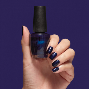 Ręka trzyma butelkę granatowej odżywki OPI Nail Envy All Night Strong, paznokcie pomalowane identycznym kolorem, tło fioletowe.