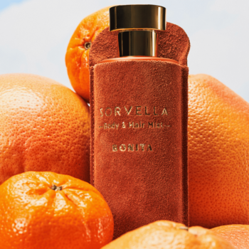 Pomarańczowa mgiełka Sorvella Bonita Body & Hair Mist wśród dojrzałych cytrusów, złota zakrętka.