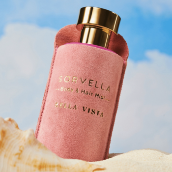 Różowa butelka Sofvella Bella Vista Body & Hair Mist w zamszowym etui, ze złotą nakrętką na piasku.