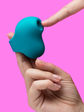 Zbliżenie: turkusowy Lovehoney Mon Ami Finger ring, stymulator ssący, palec w otworze, na różowym tle.