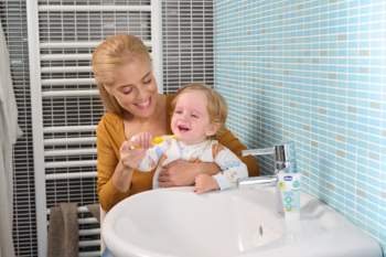 Chicco pasta do zębów jabłkowo-bananowa na umywalce, obok mama szczotkująca zęby płaczącemu dziecku.