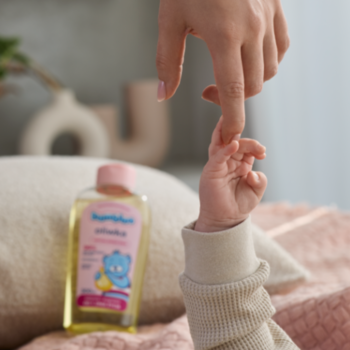 Hipoalergiczna oliwka Bambino w żółtej butelce z różową nakrętką, w tle dłoń dziecka chwytająca palec dorosłego.