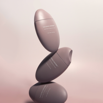 Trzy matowe butelki kremu hialuronowego z czarnym ryżem w kształcie kamyków, w odcieniu brudnego różu, ułożone w stos.