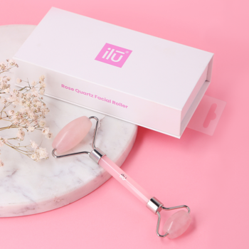 Różowy roller kwarcowy Ilū do masażu twarzy z białym pudełkiem na tle jasnego marmuru i różu.