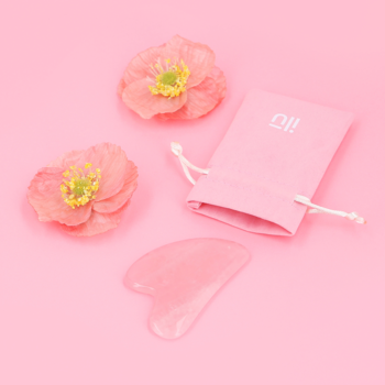 Gładki, różowy kamień Gua Sha Ilū z kwarcu do masażu, obok woreczka i dwóch kwiatów na różowym tle.