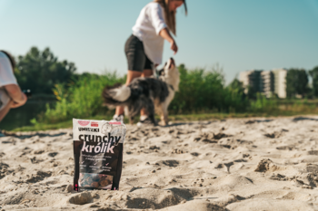 Saszetka Uniszki Crunchy Królik z miętą i pokrzywą, przysmak dla psów, na piasku. W tle rozmyty pies i człowiek.
