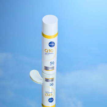 Biało-żółty NIVEA Q10 Daily UV Fluid SPF 50 stoi na odbijającej powierzchni, obok mała kropla kremu, błękitne tło.