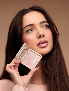 Kobieta trzyma otwarty wypiekany rozświetlacz Everybody London Pink Lights w kamieniu. Różowy produkt w kompakcie z lusterkiem.