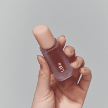 Laka Fruity Glam Tint Dewy, błyszcząca pomadka w płynie w matowej butelce, trzymana w dłoni, na jasnym tle.
