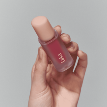 Dłoń trzyma błyszczącą pomadkę w płynie Laka Fruity Glam Tint Fresh w matowej butelce z brzoskwiniową zakrętką.