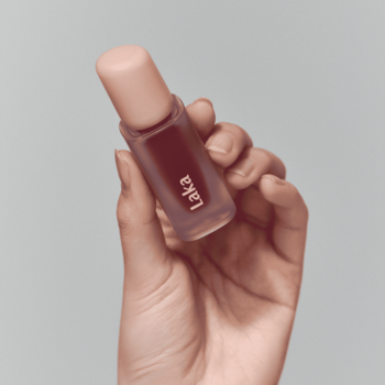 Zbliżenie dłoni trzymającej błyszczącą pomadkę Laka Fruity Glam Tint Envy: ciemny tint, brzoskwiniowa zakrętka.