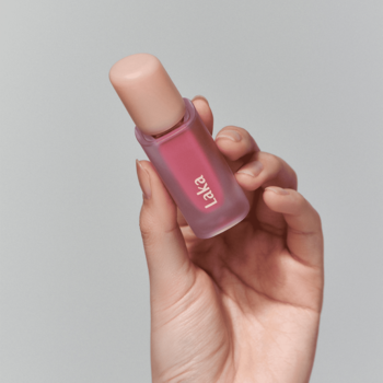 Ręka trzyma błyszczącą pomadkę Laka Fruity Glam Tint 'Dreaming' w przezroczystej butelce z różowym płynem.