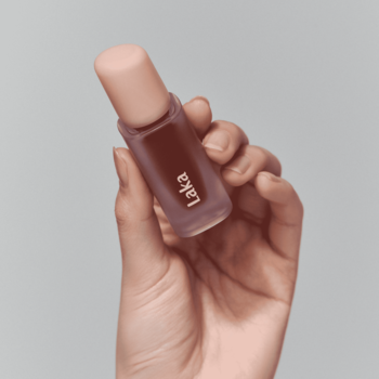 Dłoń trzyma Laka Fruity Glam Tint Caffeine Rose, błyszczyk w brązowawej butelce z różową zakrętką, pod kątem.