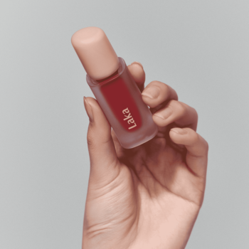 Zbliżenie na matową butelkę błyszczącej pomadki Laka Fruity Glam Tint Candid. Trzymana w dłoni, soczysta czerwień.