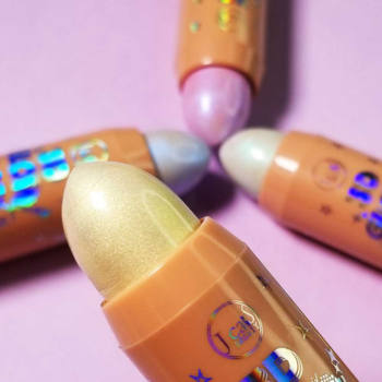 Cztery holograficzne rozświetlacze J.Cat Beauty 3D Glow Pop Holo Stick w sztyfcie, złoto, błękit, lawenda, na fioletowym tle.