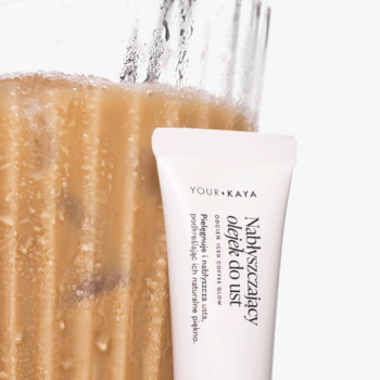 Nabłyszczający olejek do ust Your Kaya Iced Coffee Glow w białej tubce obok napoju iced coffee, widok z boku.